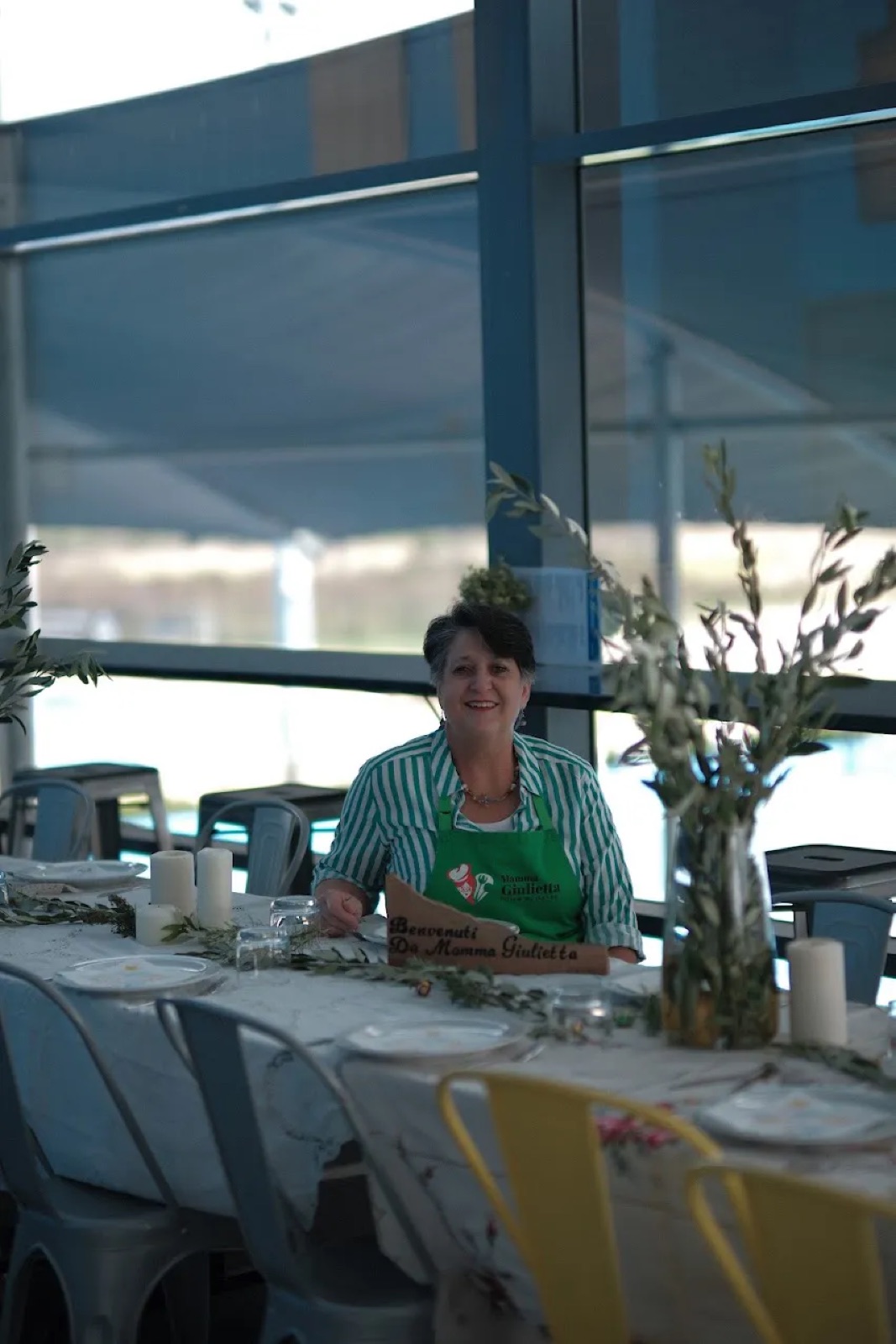 Mamma Giulietta smiling beside a long table set for lunch with a Benvenuti da Mamma Giulietta sign